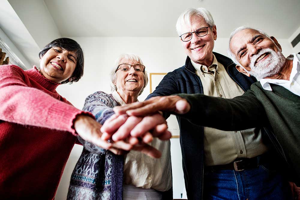 Vier lächelnde Senioren legen Hände aufeinander: Wir sind ein Team